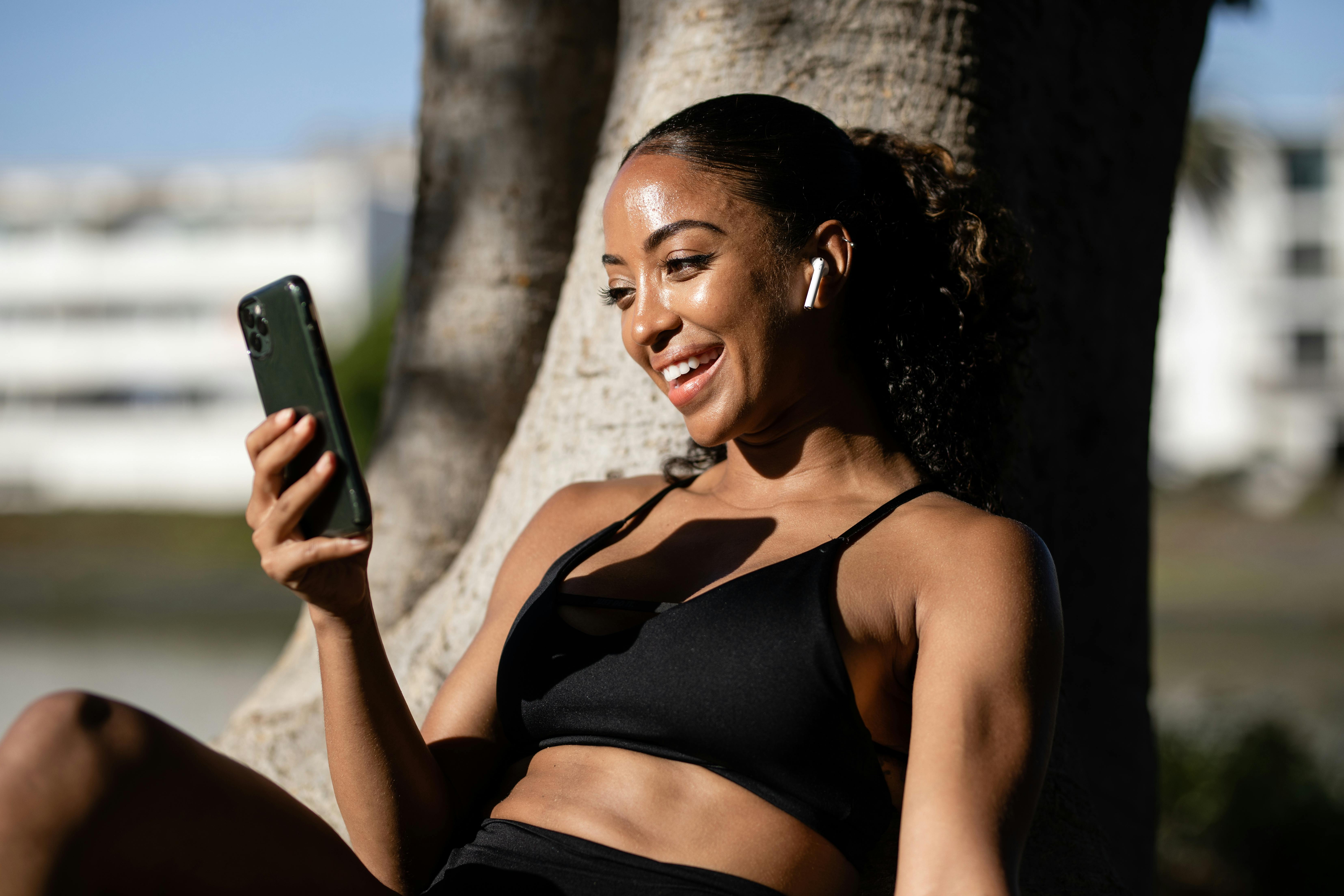 African American woman in activewear smiling during a video call outdoors with smartphone and earbuds.