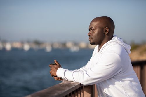 Free A man leans on a railing, overlooking a sunny waterfront while wearing a white hoodie. Stock Photo