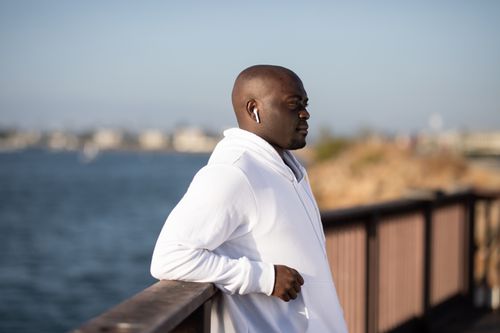Free A man in a white hoodie with earbuds, leaning on a railing by the water. Stock Photo