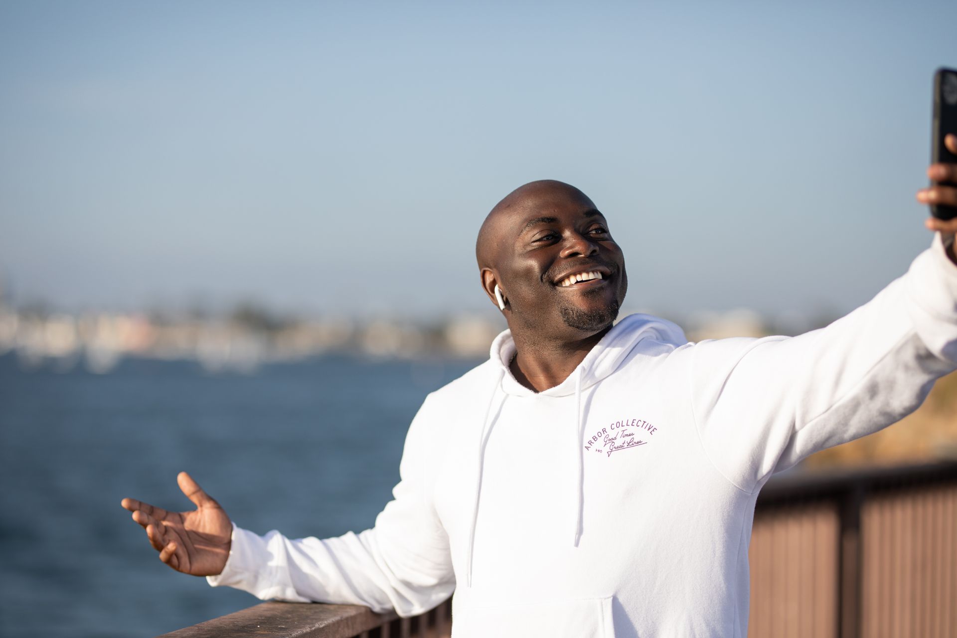 Smiling man enjoys taking a selfie by the water wearing earbuds and a hoodie.