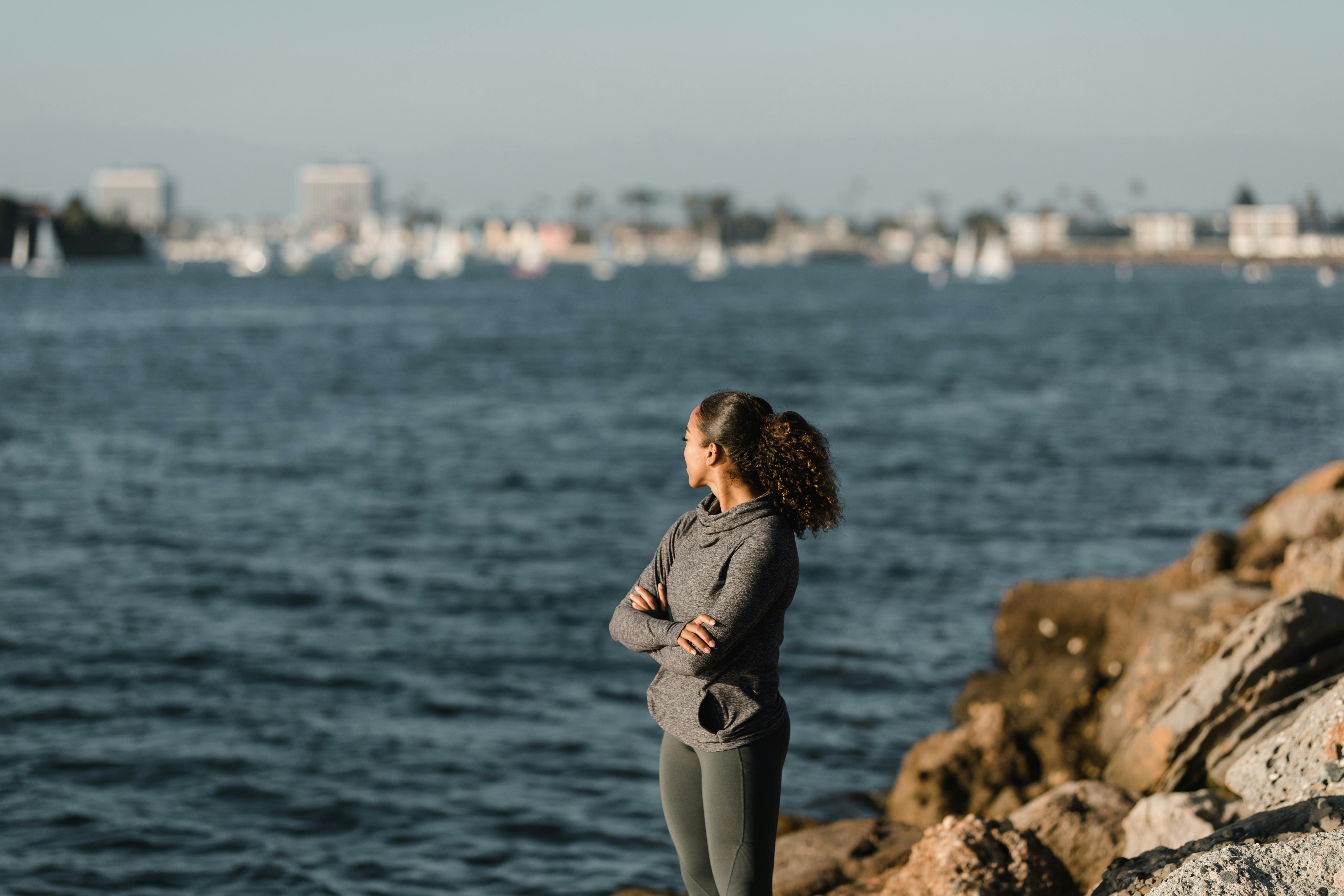 女人双臂交叉，站在岩石岸边，俯瞰着宁静的海景