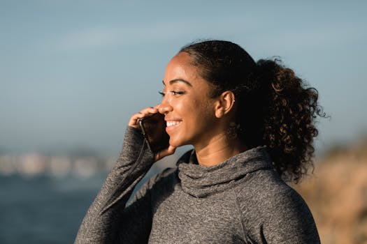 A smiling woman in a gray sweater enjoying a phone call outdoors by the sea.
