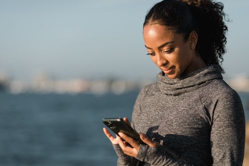 Free A woman wearing a sweater uses her smartphone near a body of water, enjoying a peaceful moment outdoors. Stock Photo