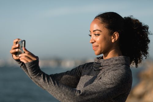Free A young woman in a sweater smiles while taking a selfie with her smartphone by the sea. Stock Photo