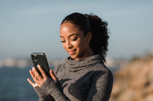 Free Smiling woman in a sweater using her smartphone by the sea in a bright day. Stock Photo