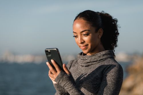 Free A woman in a gray sweater engaging with her smartphone outdoors by the water on a bright day. Stock Photo
