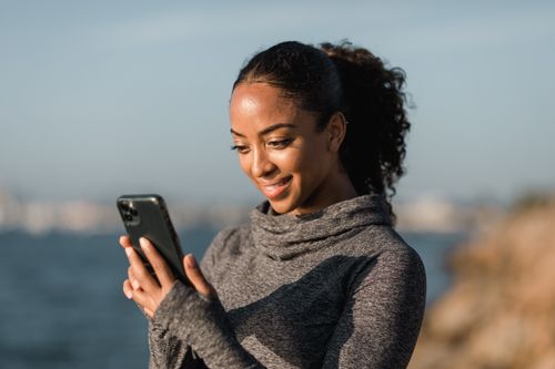 Free A smiling woman in a sweater uses her smartphone by the water on a sunny day, capturing a candid moment. Stock Photo