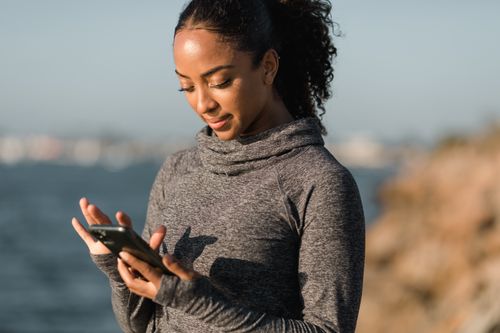 Free Young woman engaged in smartphone activity by the water, wearing a cozy grey sweater. Stock Photo