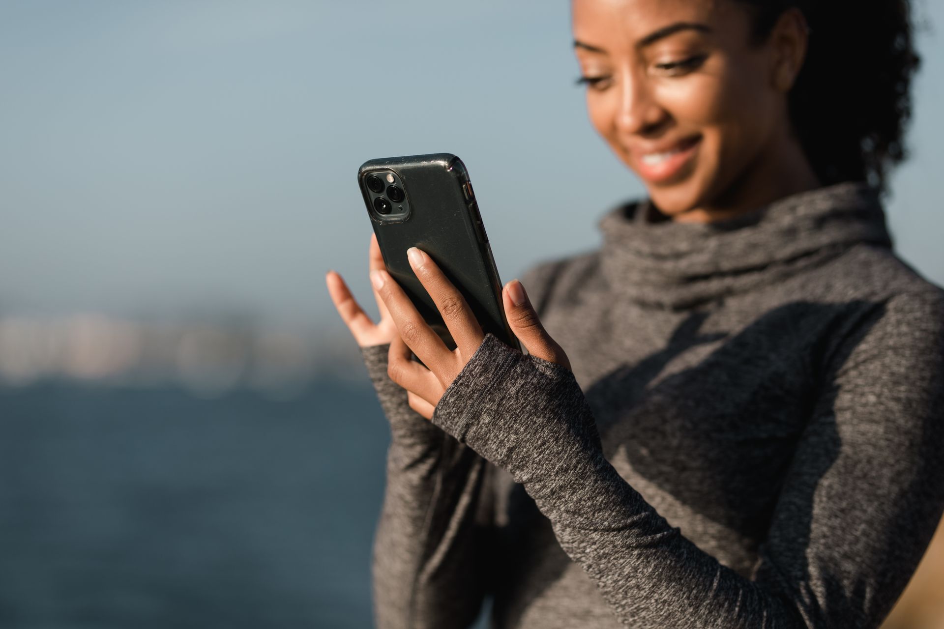 Smiling woman in a gray sweater using her smartphone outdoors near water.