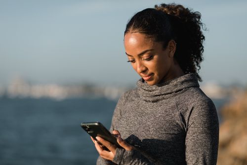 Free A young woman in a gray sweater browsing her smartphone by the seaside on a sunny day. Stock Photo
