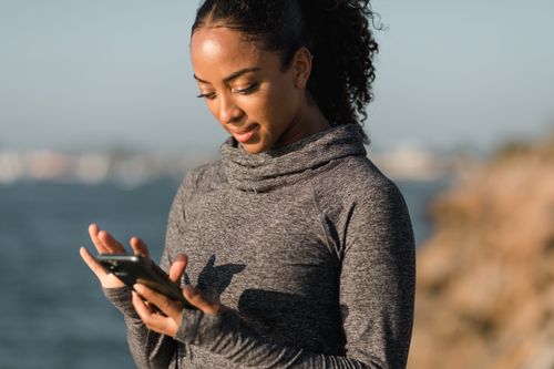 Free Woman in gray sweater using smartphone outdoors by the sea on a sunny day. Stock Photo