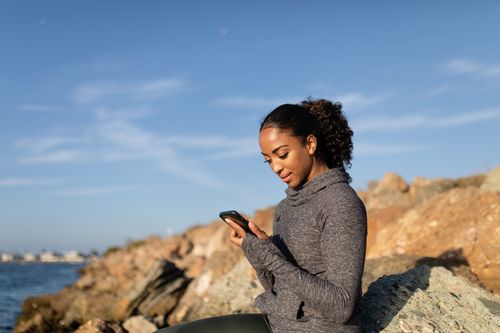 Free Young woman in sweater using smartphone on rocky beach under blue sky Stock Photo