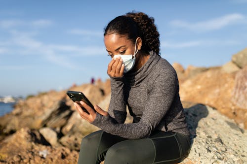 A Woman in a Face Mask Using her Cellphone while Sitting on a Rock