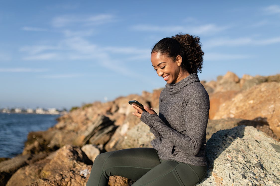 A Happy Woman Using her Cellphone while Sitting on a Rock
