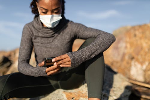 Free A Woman in a Face Mask Using her Cellphone while Sitting on a Rock Stock Photo
