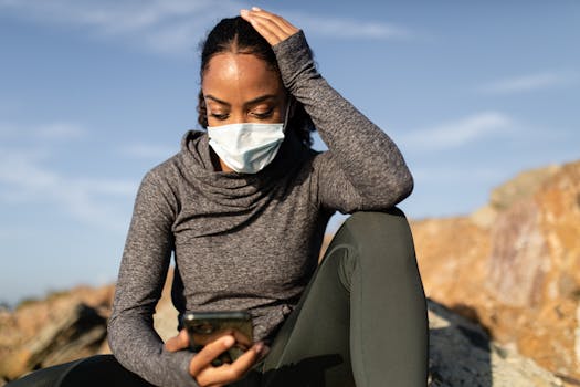 A woman wearing a face mask uses her smartphone sitting outdoors on a sunny day.