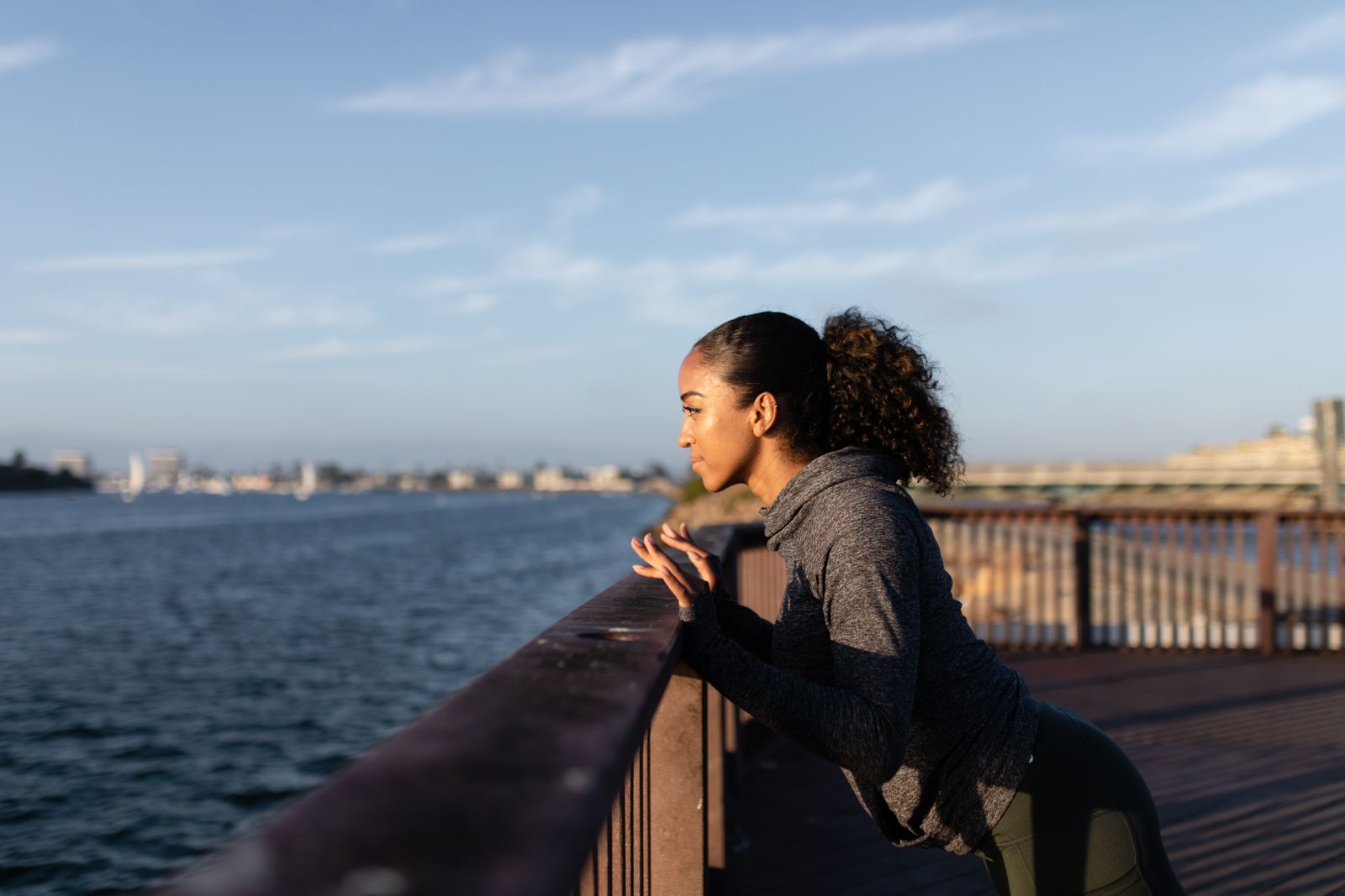 Woman in grey athletic wear leaning on a wooden pier railing overlooking the water in golden sunlight.