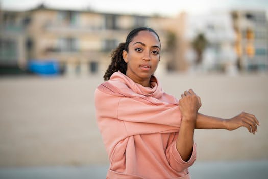 一位穿着时尚粉红色连帽衫的年轻女子，在模糊的城市背景前伸展身体