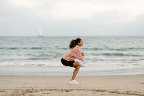Free Woman in Pink Sweater and Black Shorts Doing Yoga on Beach Shore Stock Photo