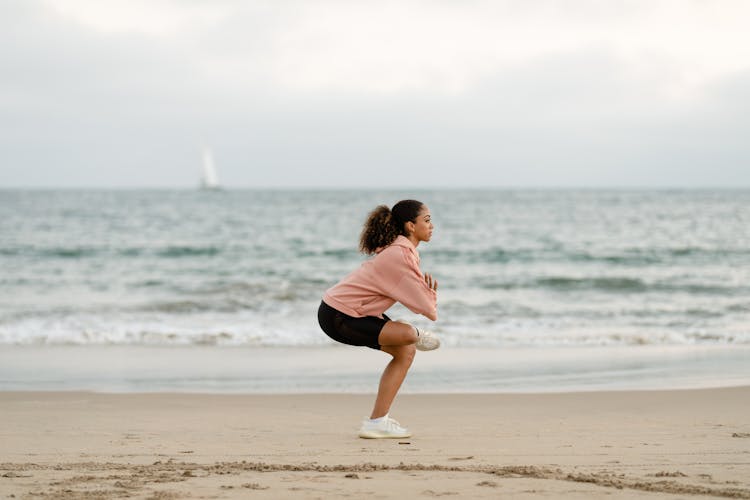 Woman In Pink Sweater And Black Shorts Doing Yoga On Beach Shore