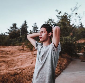 A young man stands outdoors wearing a gray shirt, hands on head in a contemplative pose.