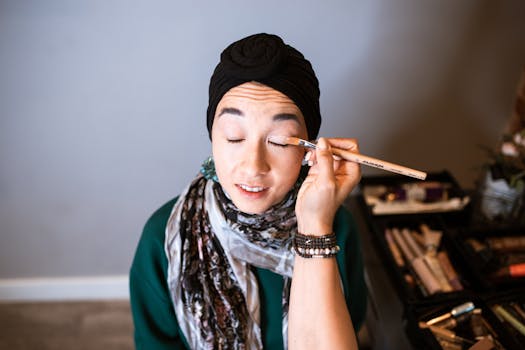 A woman in a hijab receives eye makeup application indoors, focused on beauty care.