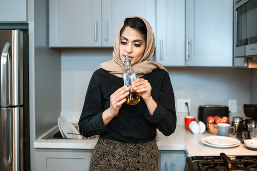 Positive stylish ethnic female in headscarf smelling aromatic olive oil in bottle while cooking dinner in contemporary light kitchen