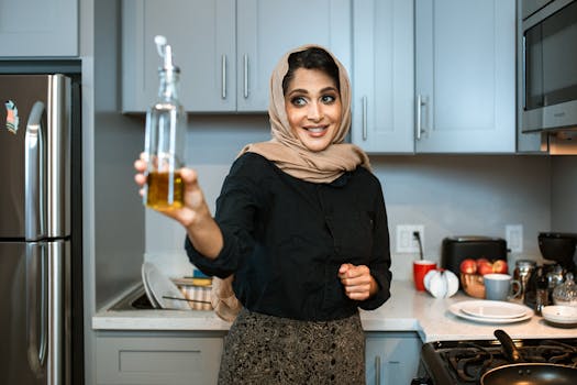 Smiling Arabic female in stylish wear and headscarf demonstrating glass bottle of olive or sunflower oil while cooking dinner in modern light kitchen