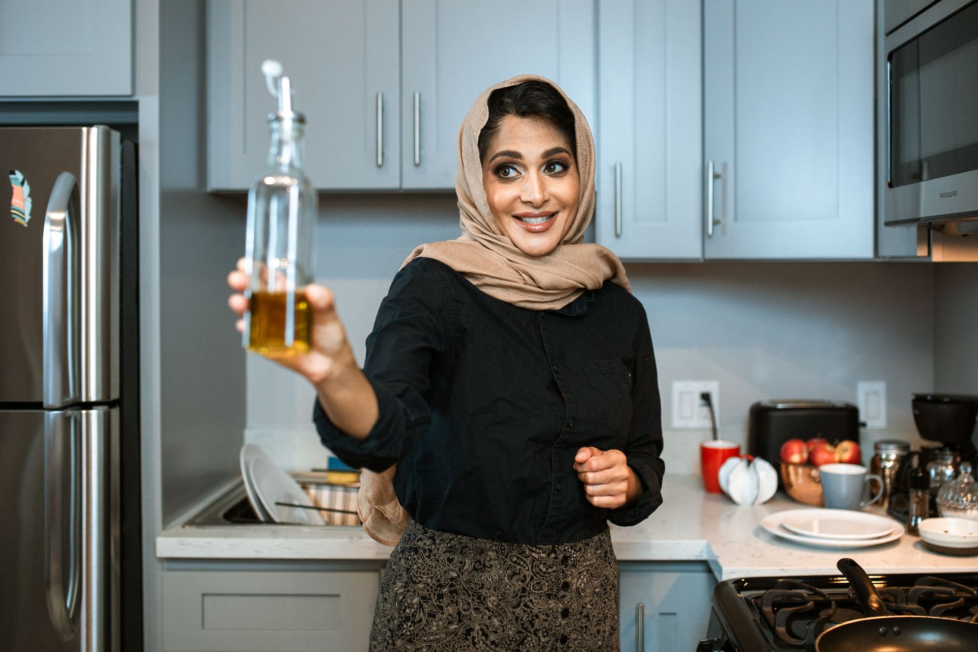 Smiling Arabic female in stylish wear and headscarf demonstrating glass bottle of olive or sunflower oil while cooking dinner in modern light kitchen