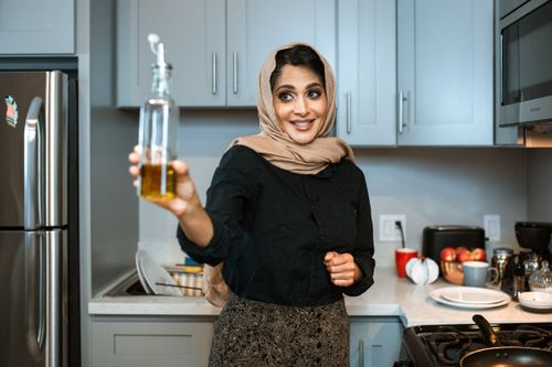 Free Smiling Arabic female in stylish wear and headscarf demonstrating glass bottle of olive or sunflower oil while cooking dinner in modern light kitchen Stock Photo