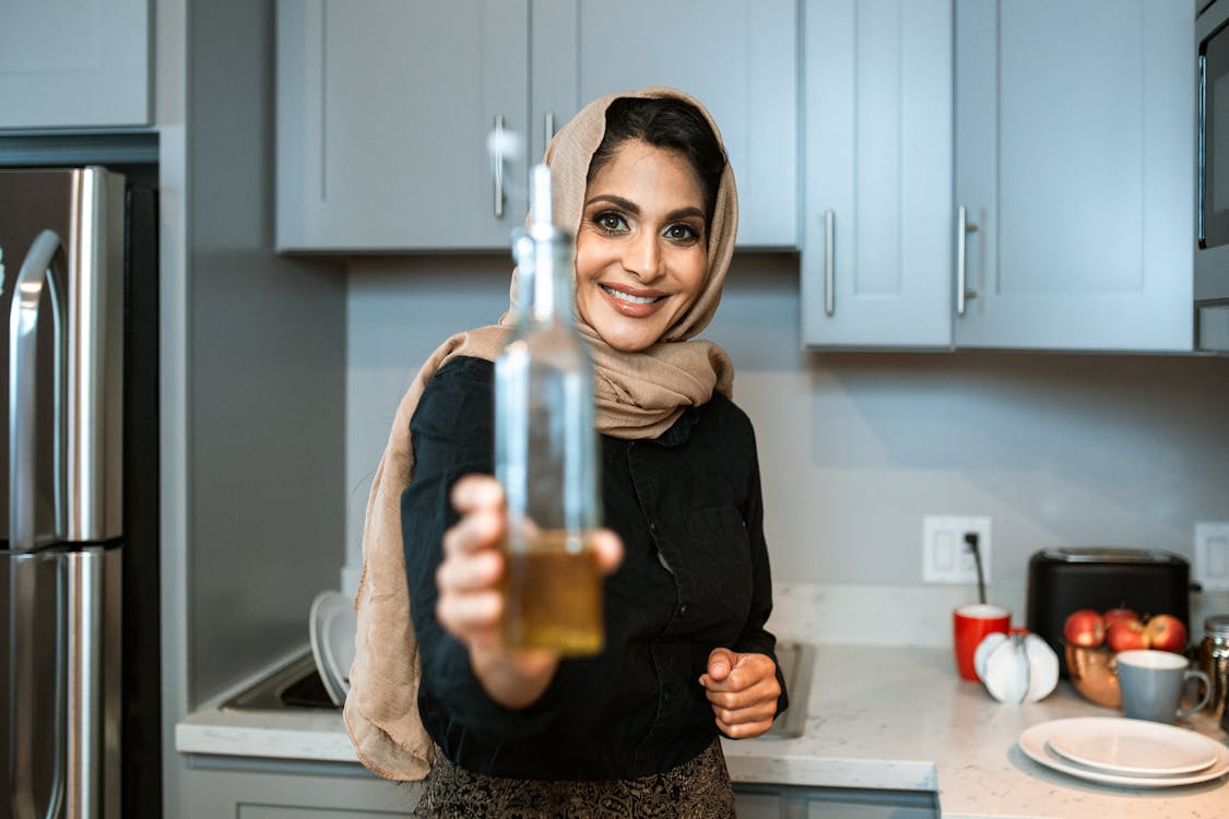 Happy ethnic female in headscarf showing bottle of organic olive oil and looking at camera with smile while cooking in contemporary kitchen