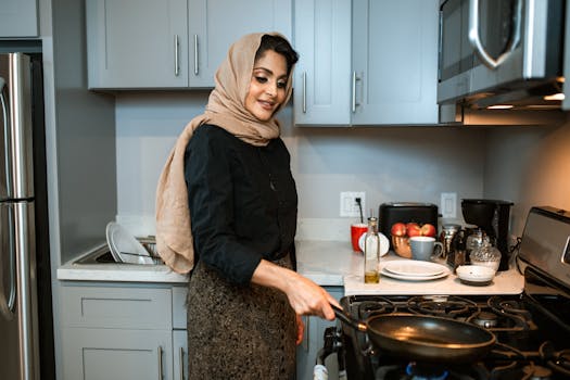 A cheerful woman wearing a headscarf cooks a meal in a contemporary kitchen.