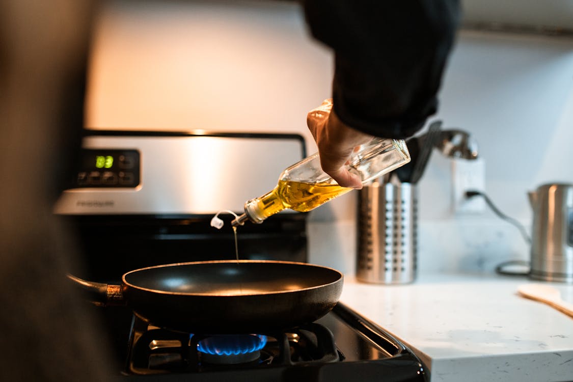 Free Back view crop unrecognizable person pouring olive or sunflower oil into frying pan placed on stove in domestic kitchen Stock Photo