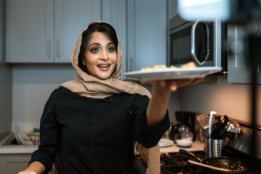 A joyful woman wearing a hijab presenting a freshly cooked dish in a contemporary kitchen setting.
