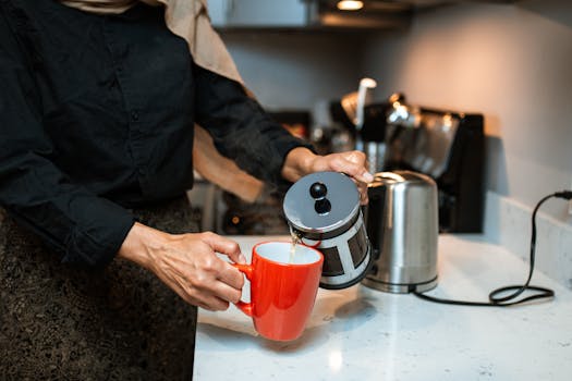 A woman uses a French press to pour coffee into a red ceramic mug on a modern kitchen countertop.