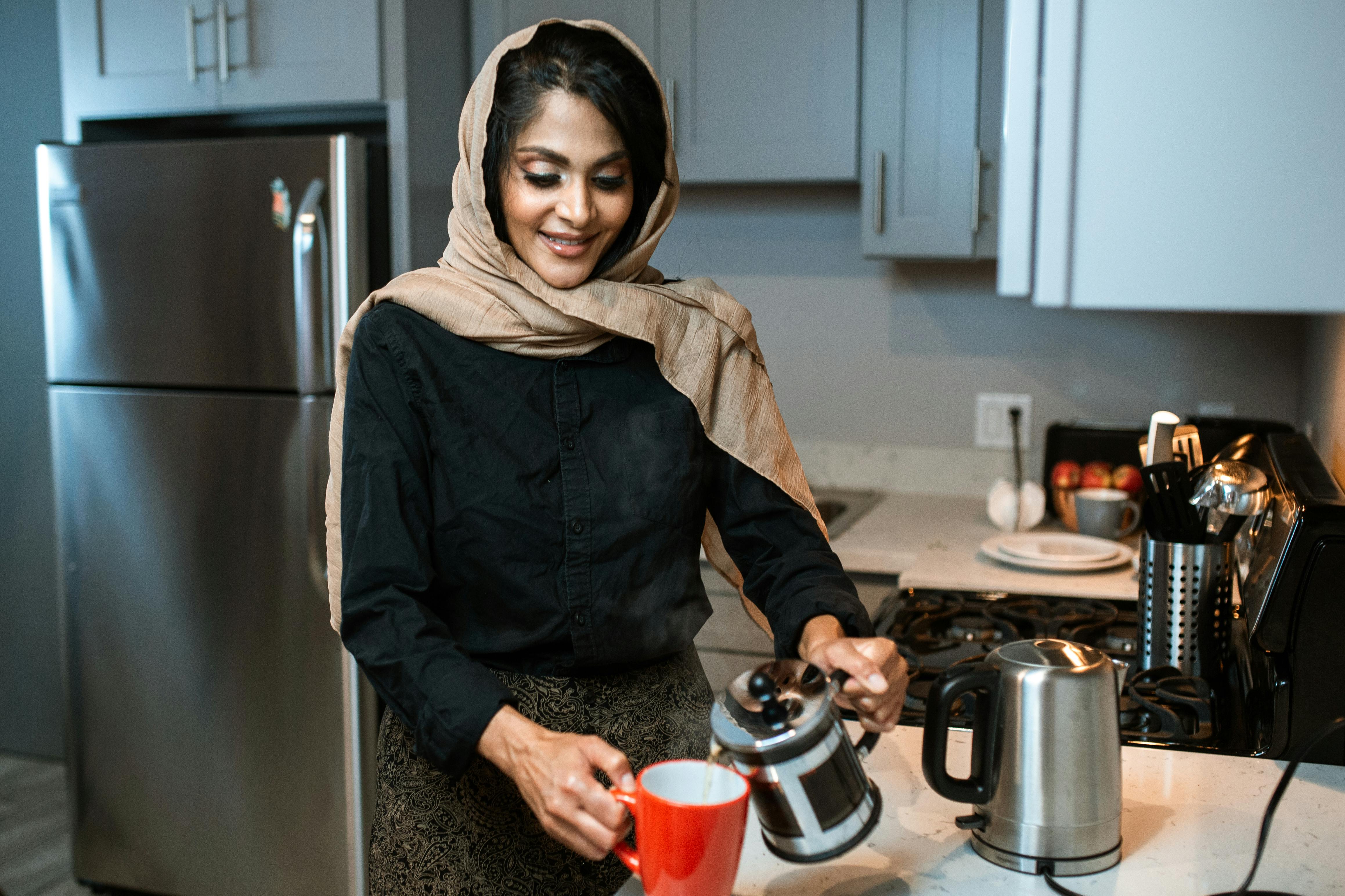 A woman wearing a hijab smiles while pouring coffee in a modern kitchen setting.