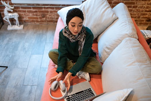 Young woman in hijab sitting on couch with laptop and headphones, focused on remote work.
