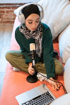 A woman wearing headphones records podcast using a condenser microphone and laptop.