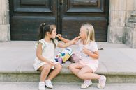 Two young girls enjoying snacks together on stone steps in a warm outdoor setting.