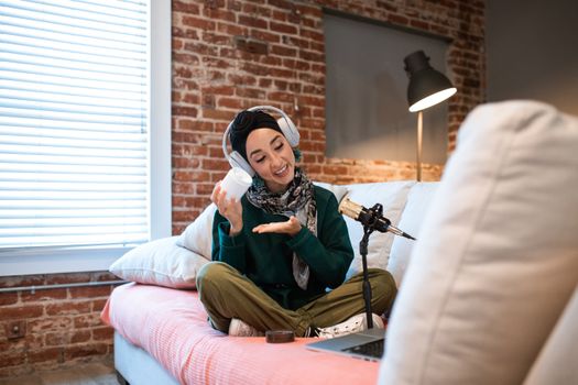 Woman podcasting at home, wearing headphones and scarf, with microphone and laptop.