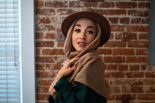 Free Stylish woman wearing a hijab and brown fedora posing indoors with a rustic brick wall background. Stock Photo