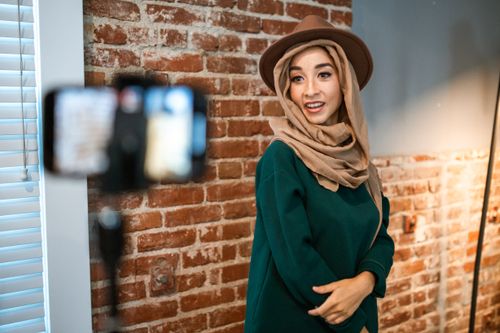 Free Fashion blogger wearing hijab and fedora hat poses indoors against a brick wall, captured during a vlogging session. Stock Photo
