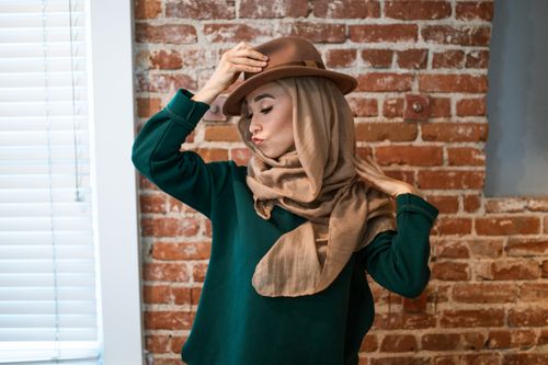 Free Stylish woman in hijab and fedora posing against a brick wall indoors. Stock Photo