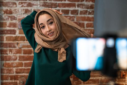 Free Woman in hijab posing in front of camera, creating vlog content indoors against a brick wall. Stock Photo