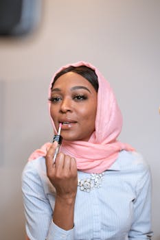 Portrait of a stylish Muslim woman in a pink headscarf applying lip gloss indoors.