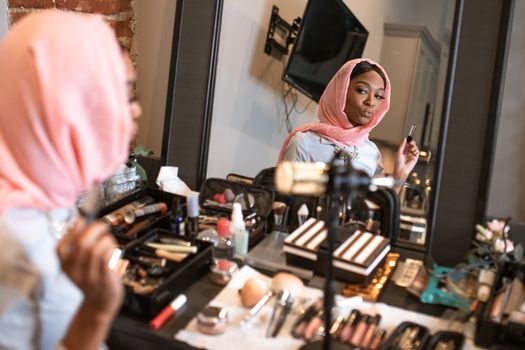 Fashionable woman in hijab applying makeup reflects in a mirror.