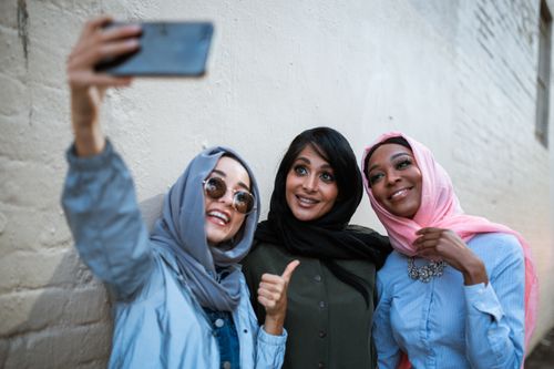 Free Three women in hijabs smiling and taking a selfie outdoors on a brick wall background. Stock Photo