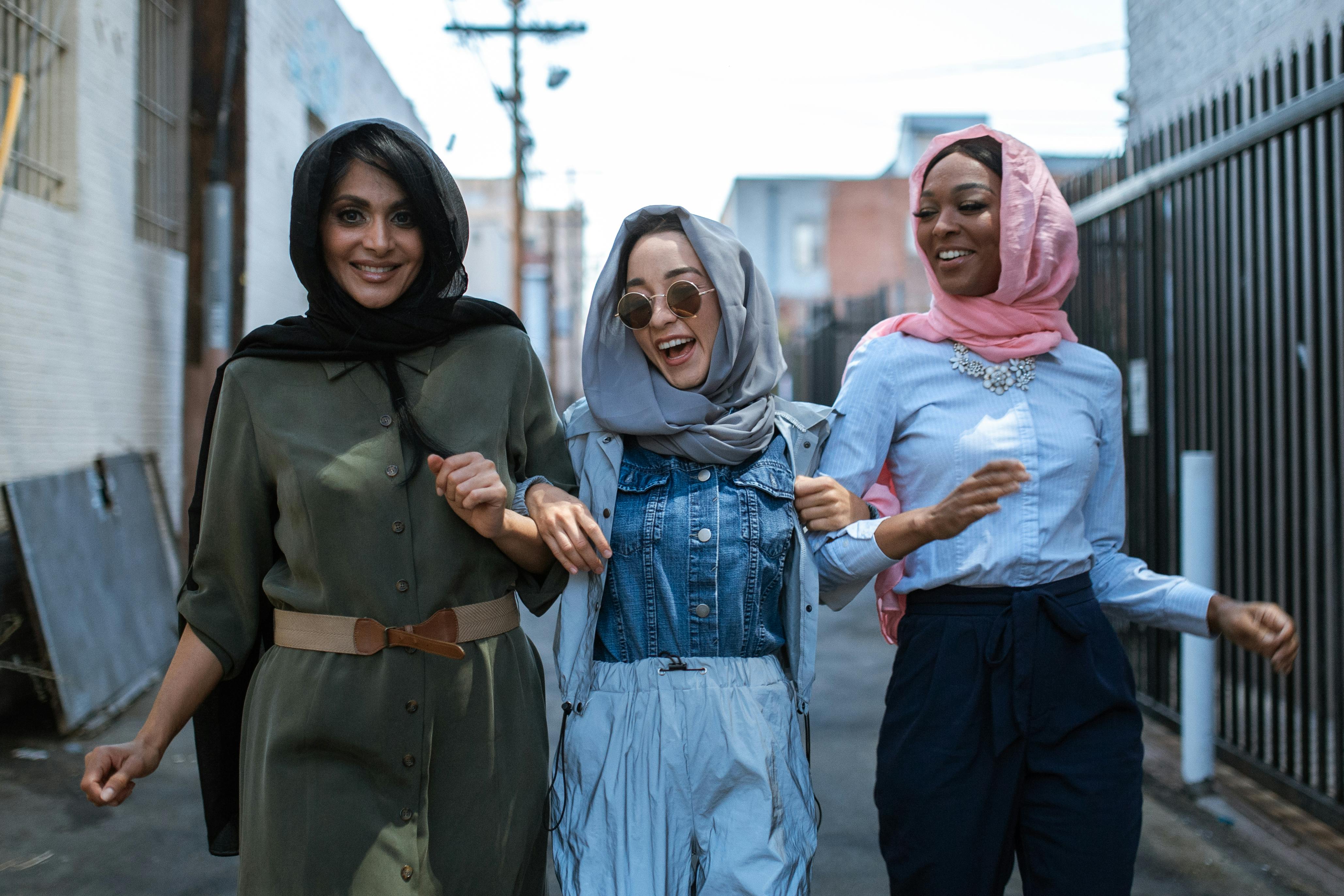 Delighted multiracial Muslim females wearing trendy outfits holding arms and walking together on narrow street and laughing