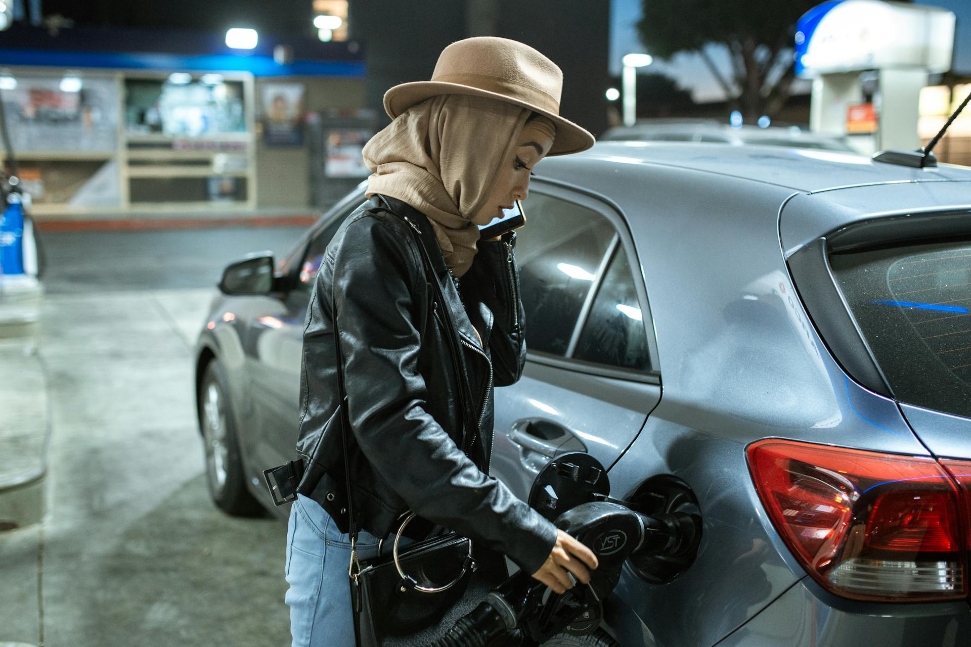 A woman in a hijab and hat refuels her car at a gas station, using a fuel pump.