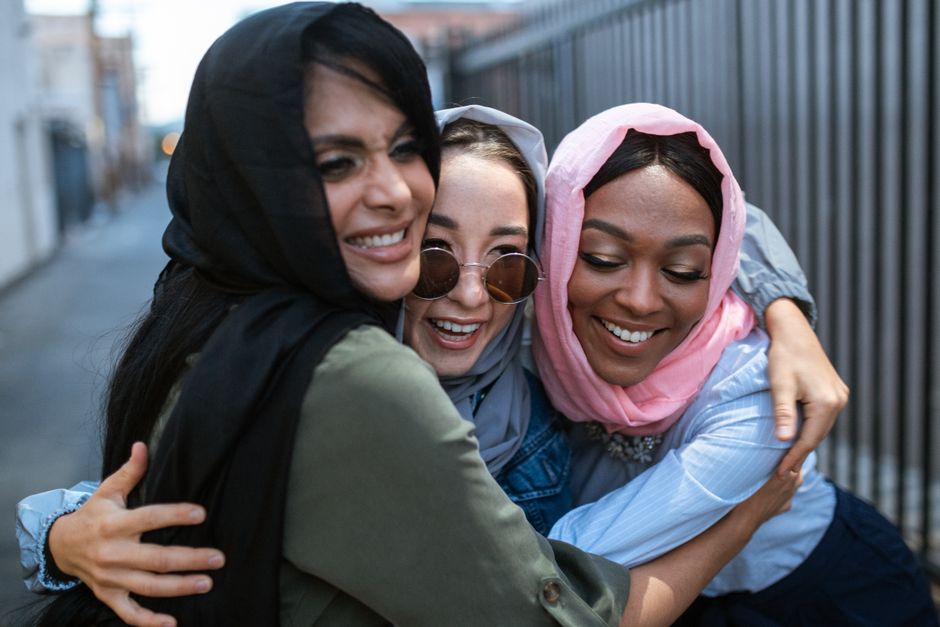 Three diverse women in colorful hijabs share a joyful embrace in an urban setting under daylight.